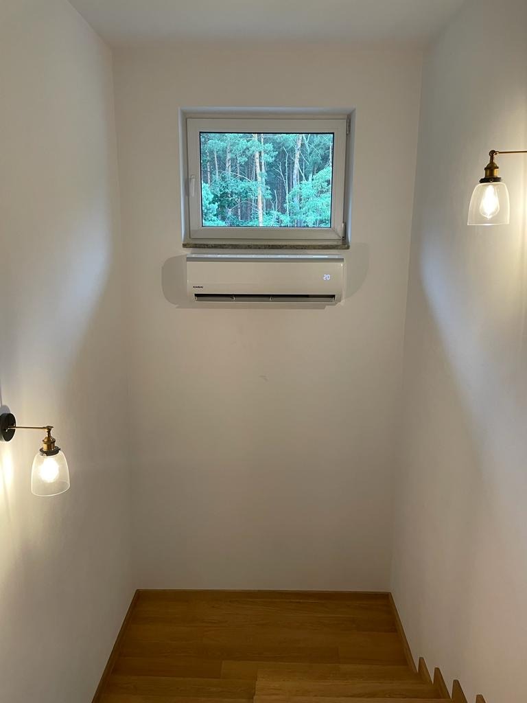 Biała klimatyzacja ścienna pod oknem z widokiem na las, oświetlona przez dwie lampy ścienne z żarówkami Edisona, na tle białych ścian i drewnianych schodów.