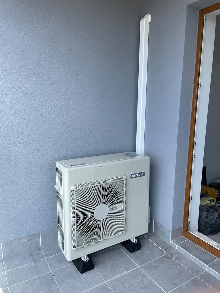 Biała jednostka zewnętrzna klimatyzacji Hitachi, zamontowana na czarnych podkładkach, na tle szarej ściany i płytek. Widoczna rura odprowadzająca skropliny.