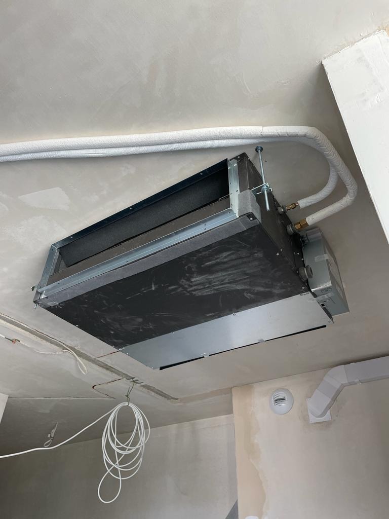 Sufitowa jednostka klimatyzacji kanałowej w surowym wnętrzu, widoczne białe rury i przewody, szara ściana w tle. Montaż klimatyzacji w trakcie prac wykończeniowych.