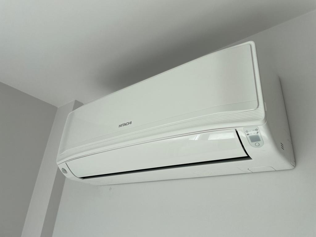Biała klimatyzacja Hitachi zamontowana na ścianie w jasnym pomieszczeniu. Widoczny panel sterowania i logo producenta. Minimalistyczny design.