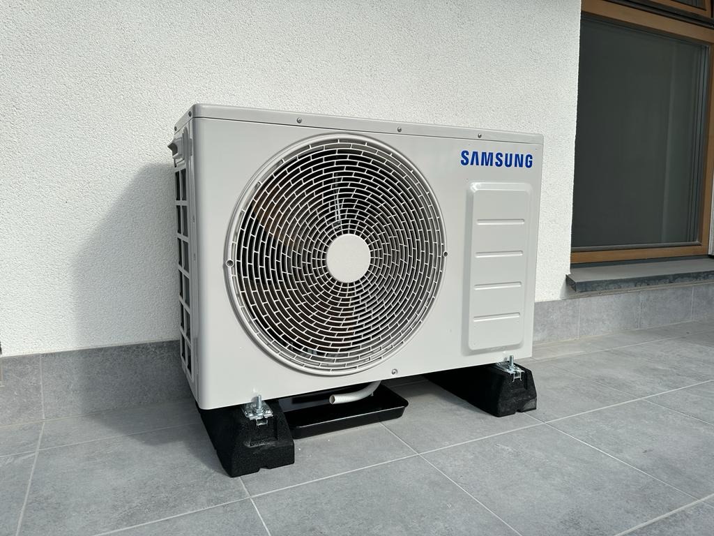 Biała jednostka zewnętrzna klimatyzacji Samsung na czarnych podkładkach antywibracyjnych, ustawiona na szarych płytkach, z oknem w tle.