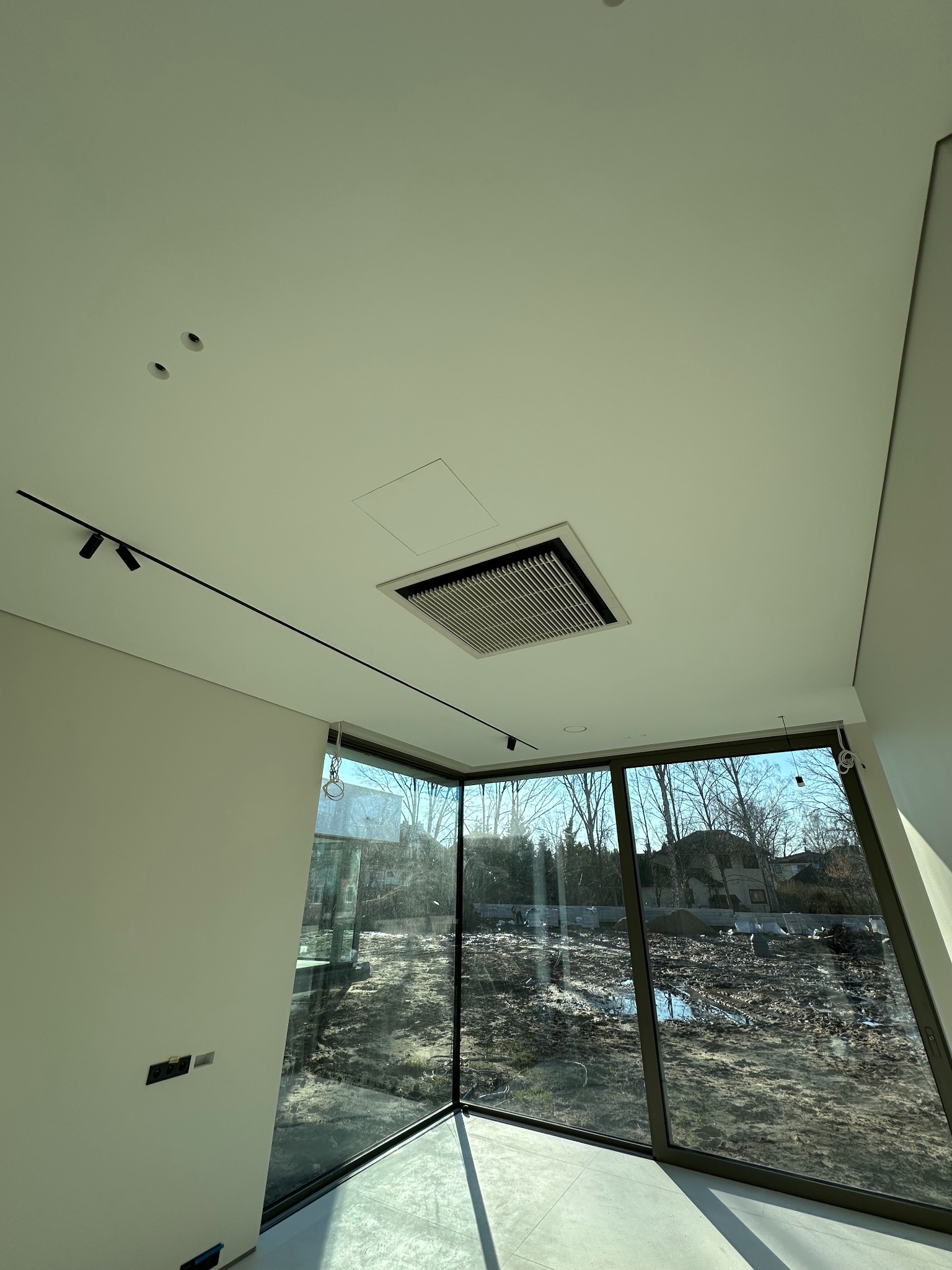 Nowoczesne wnętrze z dużym oknem i wbudowaną w sufit kratką wentylacyjną. Widoczne oświetlenie szynowe i minimalistyczne wykończenie ścian.