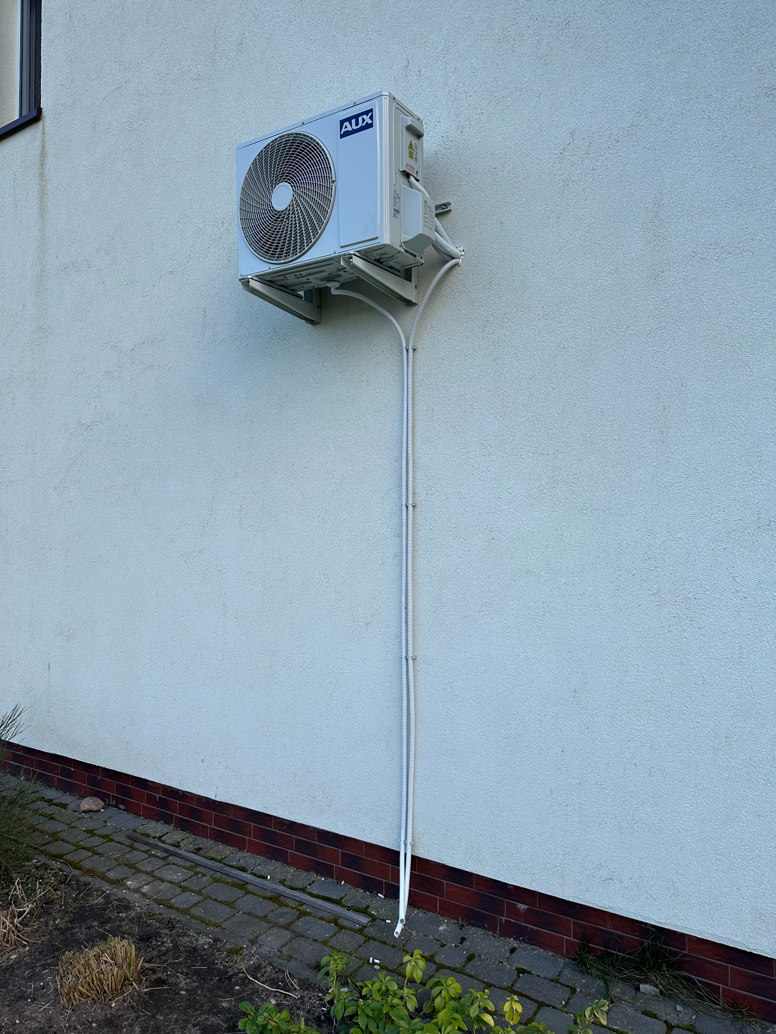 Biała jednostka zewnętrzna klimatyzacji AUX zamontowana na elewacji budynku z widocznymi przewodami w białych osłonach. Ujęcie z dołu, z fragmentem chodnika i roślinności.