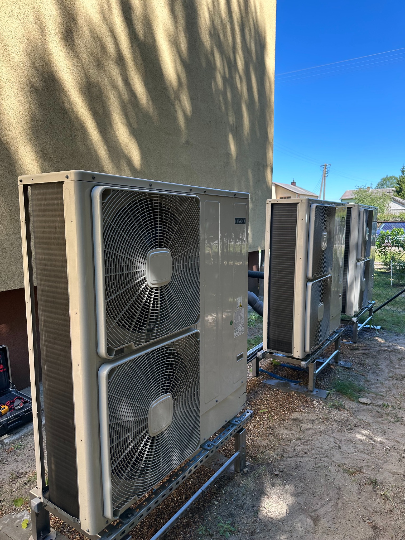 Rząd zamontowanych, zewnętrznych jednostek klimatyzacji Hitachi na metalowych konstrukcjach, ustawionych wzdłuż ściany budynku w słoneczny dzień. Widoczne narzędzia w tle.