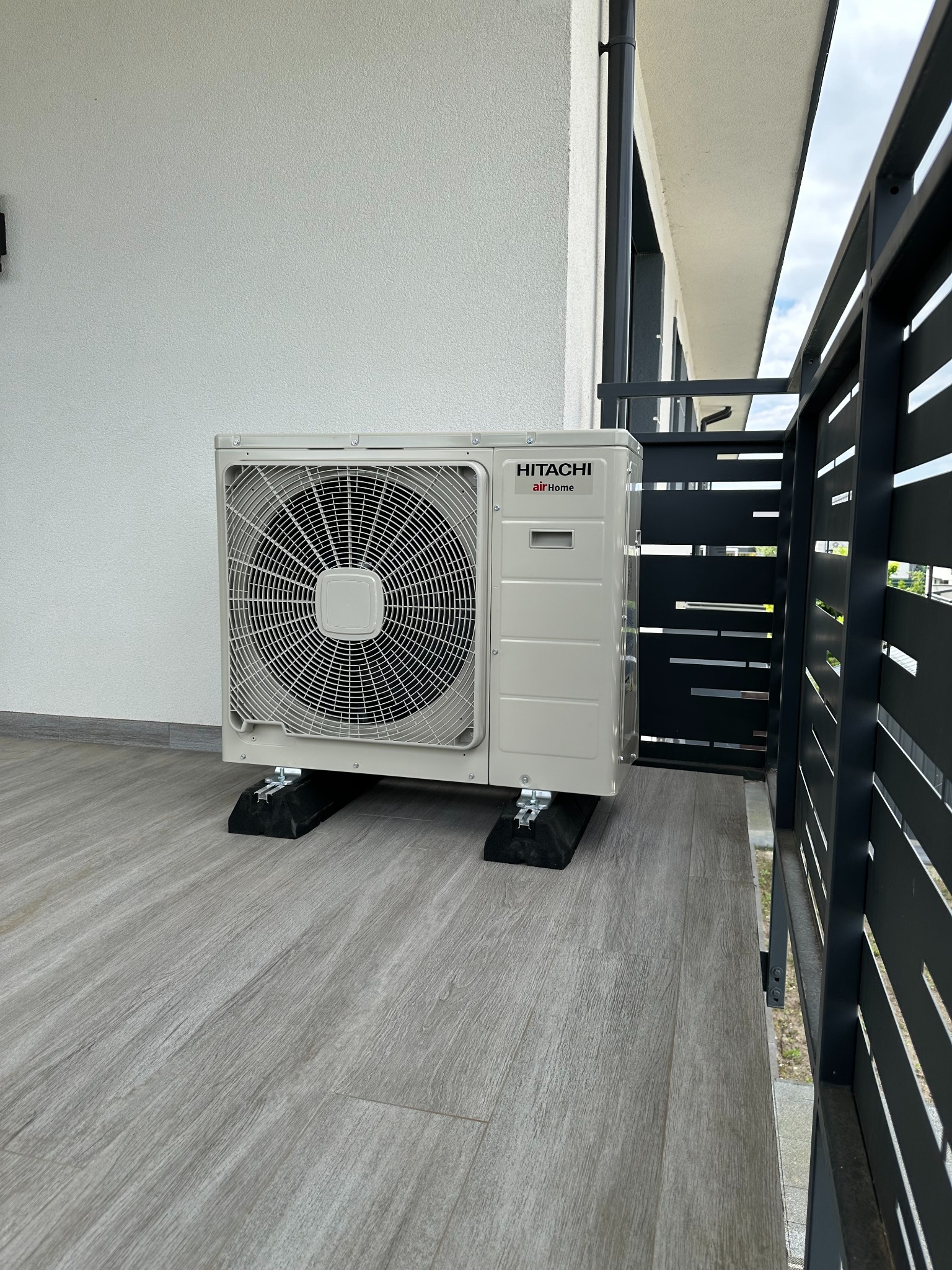 Zewnętrzna jednostka klimatyzacji Hitachi airHome na balkonie z szarą podłogą, zamontowana na czarnych podkładkach antywibracyjnych, widok z boku, nowoczesne ogrodzenie.