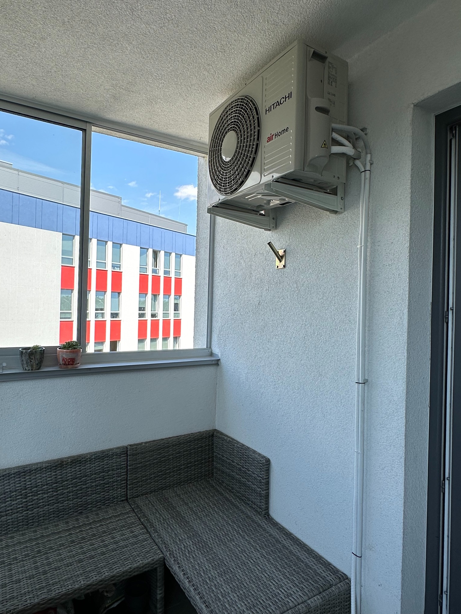 Klimatyzator Hitachi Air Home zamontowany na balkonie, widoczne rury instalacyjne prowadzone wzdłuż ściany. W tle budynek z czerwono-białą fasadą.