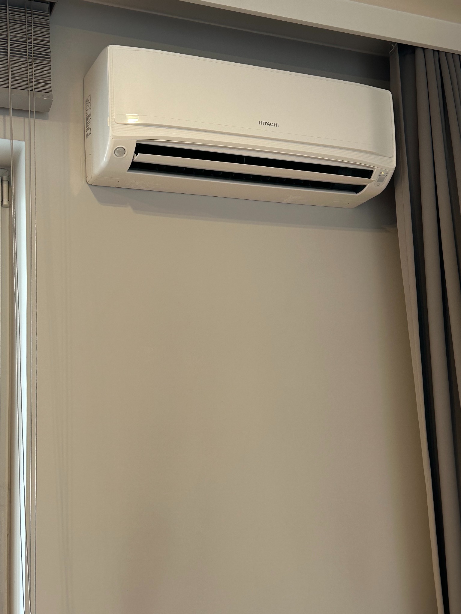 Biały klimatyzator Hitachi zamontowany na beżowej ścianie w pomieszczeniu z zasłonami i żaluzjami. Widoczne detale urządzenia i otoczenia.