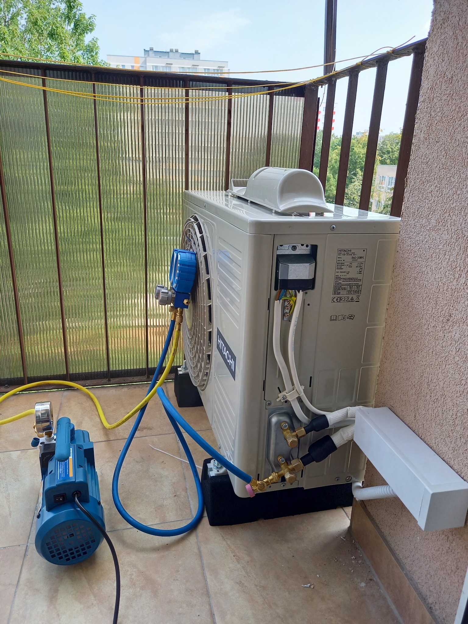 Jednostka zewnętrzna klimatyzacji Hitachi na balkonie, podłączona do manometrów serwisowych i pompy próżniowej podczas serwisu.