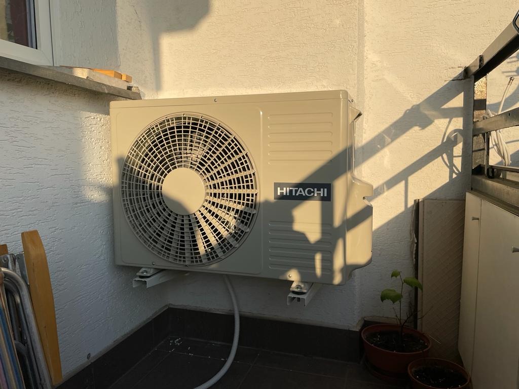 Zewnętrzna jednostka klimatyzacji Hitachi zamontowana na balkonie, oświetlona popołudniowym słońcem, z widocznymi cieniami na ścianie i podłodze.