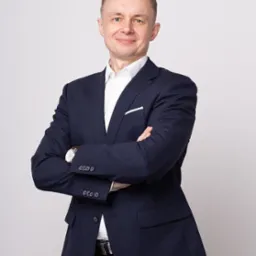 Portret uśmiechniętego mężczyzny w ciemnym garniturze i białej koszuli, z zegarkiem na nadgarstku, stojącego na jasnoszarym tle z założonymi rękami.