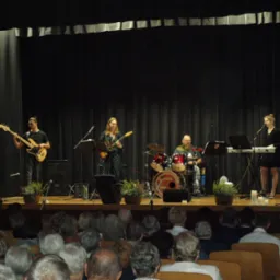 Zespół muzyczny gra koncert na scenie przed publicznością; wokalistka śpiewa do mikrofonu, gitarzysta gra na gitarze basowej, gitarzystka na gitarze elektrycznej, a perkusista na perkusji.