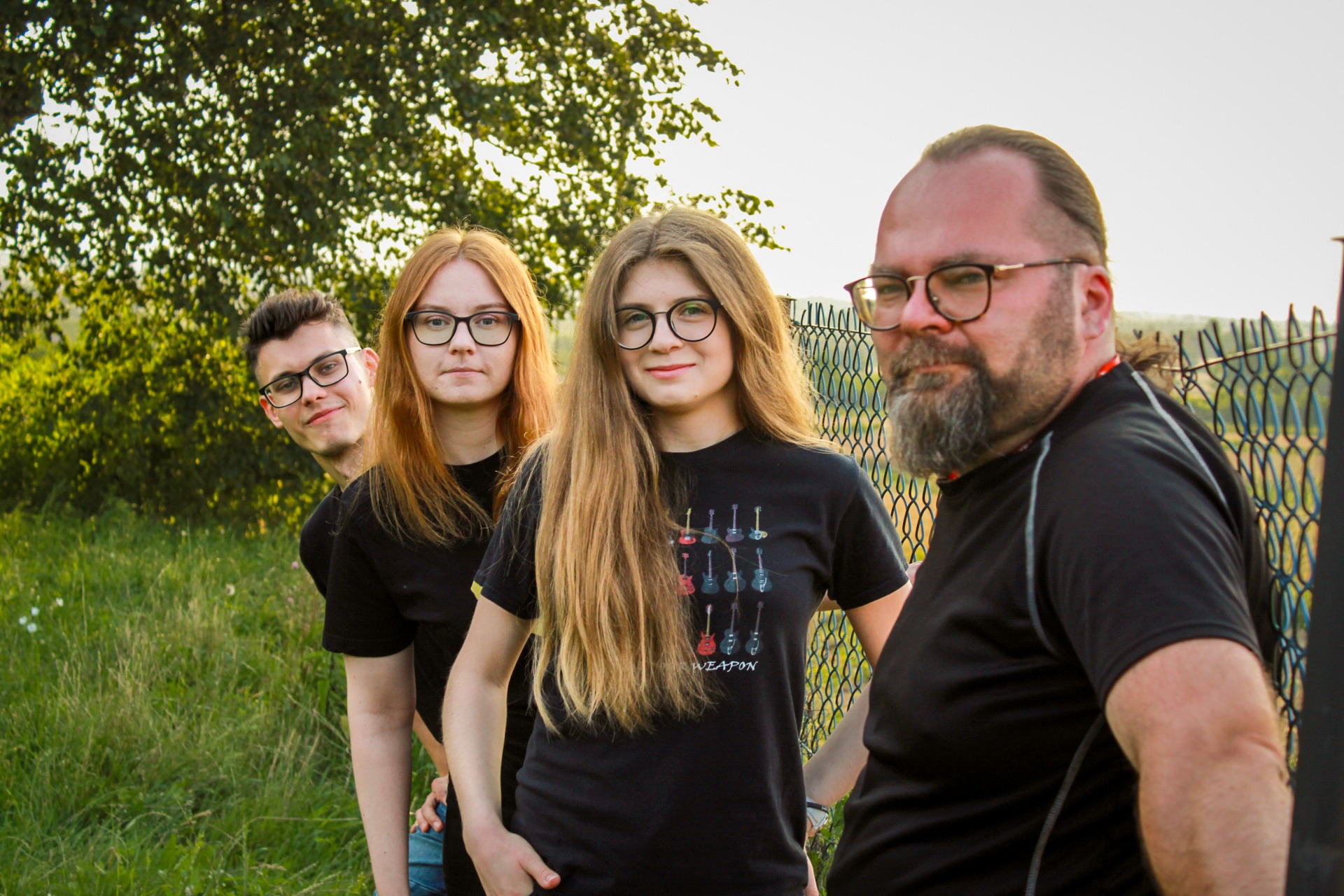 Czteroosobowa grupa, prawdopodobnie muzyczna, pozuje na zewnątrz, na pierwszym planie dziewczyna w czarnej koszulce z grafiką gitar, za nią reszta zespołu, w tle drzewa i ogrodzenie.
