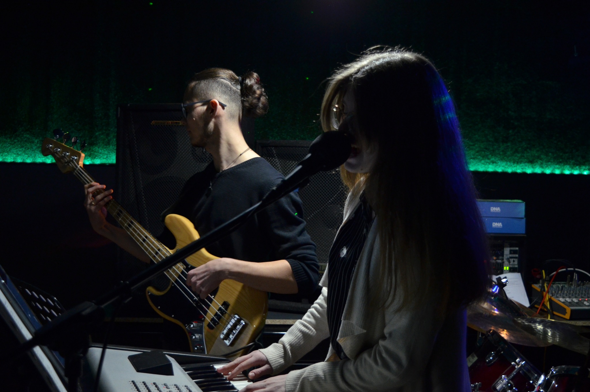 Występ na scenie: basista z gitarą basową i klawiszowiec z mikrofonem podczas koncertu, w tle zielone oświetlenie sceniczne.