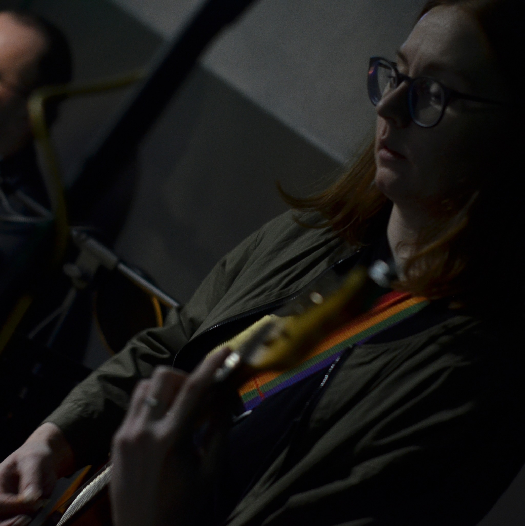 Kobieta w okularach gra na gitarze elektrycznej z tęczowym paskiem w ciemnym otoczeniu.