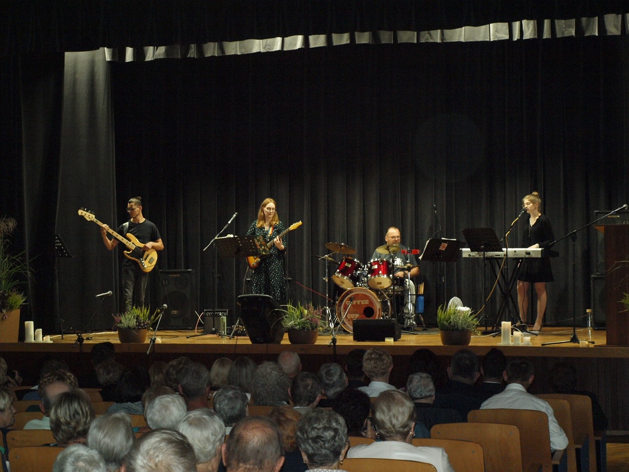 Zespół muzyczny gra koncert na scenie przed publicznością; wokalistka śpiewa do mikrofonu, gitarzysta gra na gitarze basowej, gitarzystka na gitarze elektrycznej, a perkusista na perkusji.
