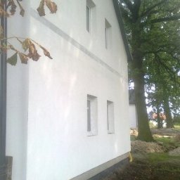 KOMPLEX-DOM Robert Łach - Boczny widok otynkowanej na biało ściany domu z czterema oknami, widoczna szara linia dekoracyjna, drzewo w tle, świeżo obsypana ziemia wokół fundamentów.