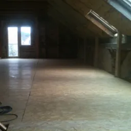 Płyty OSB ułożone na podłodze poddasza w trakcie prac wykończeniowych, widoczne okno dachowe i fragment kabla elektrycznego w rogu.