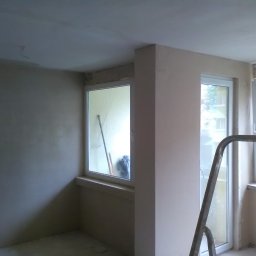 KOMPLEX-DOM Robert Łach - Wnętrze w trakcie szpachlowania ścian, widoczna drabina oparta o ścianę, okna z widokiem na zewnątrz oraz częściowo wyszpachlowana ściana z pozostałościami starej warstwy.