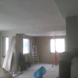 KOMPLEX-DOM Robert Łach - Wnętrze w trakcie prac wykończeniowych, ściany i sufit po szpachlowaniu, widoczne okna, drabina, podnośnik, materiały budowlane i instalacja kominowa.