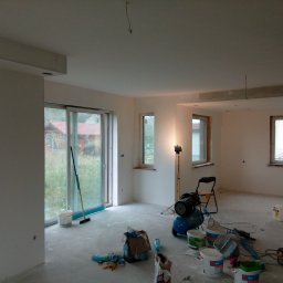 KOMPLEX-DOM Robert Łach - Pokój w trakcie prac wykończeniowych: świeżo wyszpachlowane ściany, narzędzia malarskie, kompresor, drabina i wiaderka z farbą na podłodze, w tle widok na ogród przez przesuwne okno.