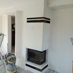 KOMPLEX-DOM Robert Łach - Nowoczesny kominek w trakcie wykończenia, widoczny odkurzacz przemysłowy Karcher, ślady po szpachlowaniu na ścianach i podłodze, surowy stan wnętrza.