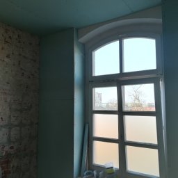 KOMPLEX-DOM Robert Łach - Montaż płyt gipsowo-kartonowych na ścianach i suficie w pokoju z oknem o łukowatym kształcie, widoczny fragment surowej ceglanej ściany i narzędzia na parapecie.