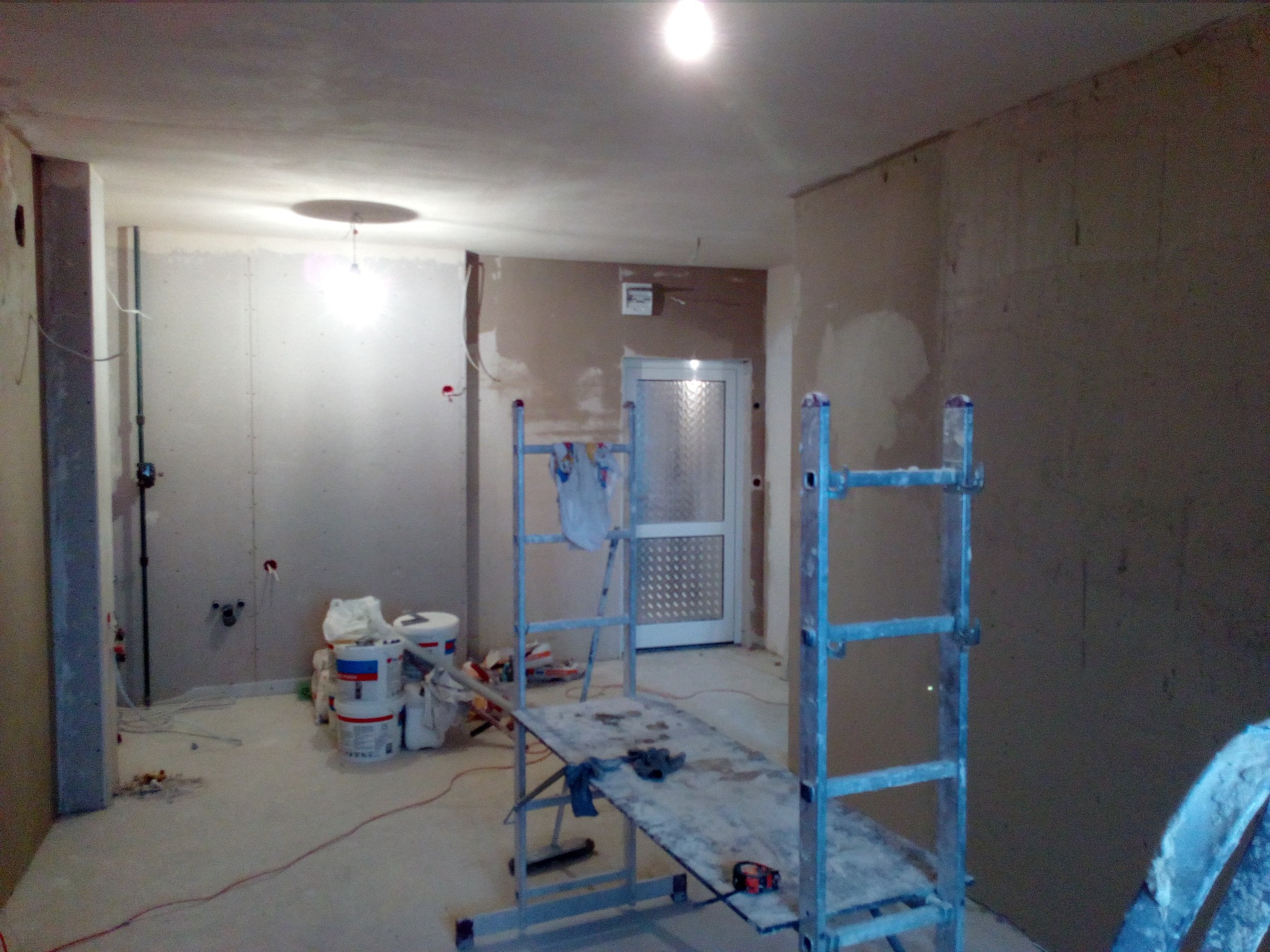 Wnętrze w trakcie prac wykończeniowych: ściany przygotowane do malowania, widoczne puszki z farbą, rozstawiony rusztowanie z narzędziami, surowy beton na podłodze.