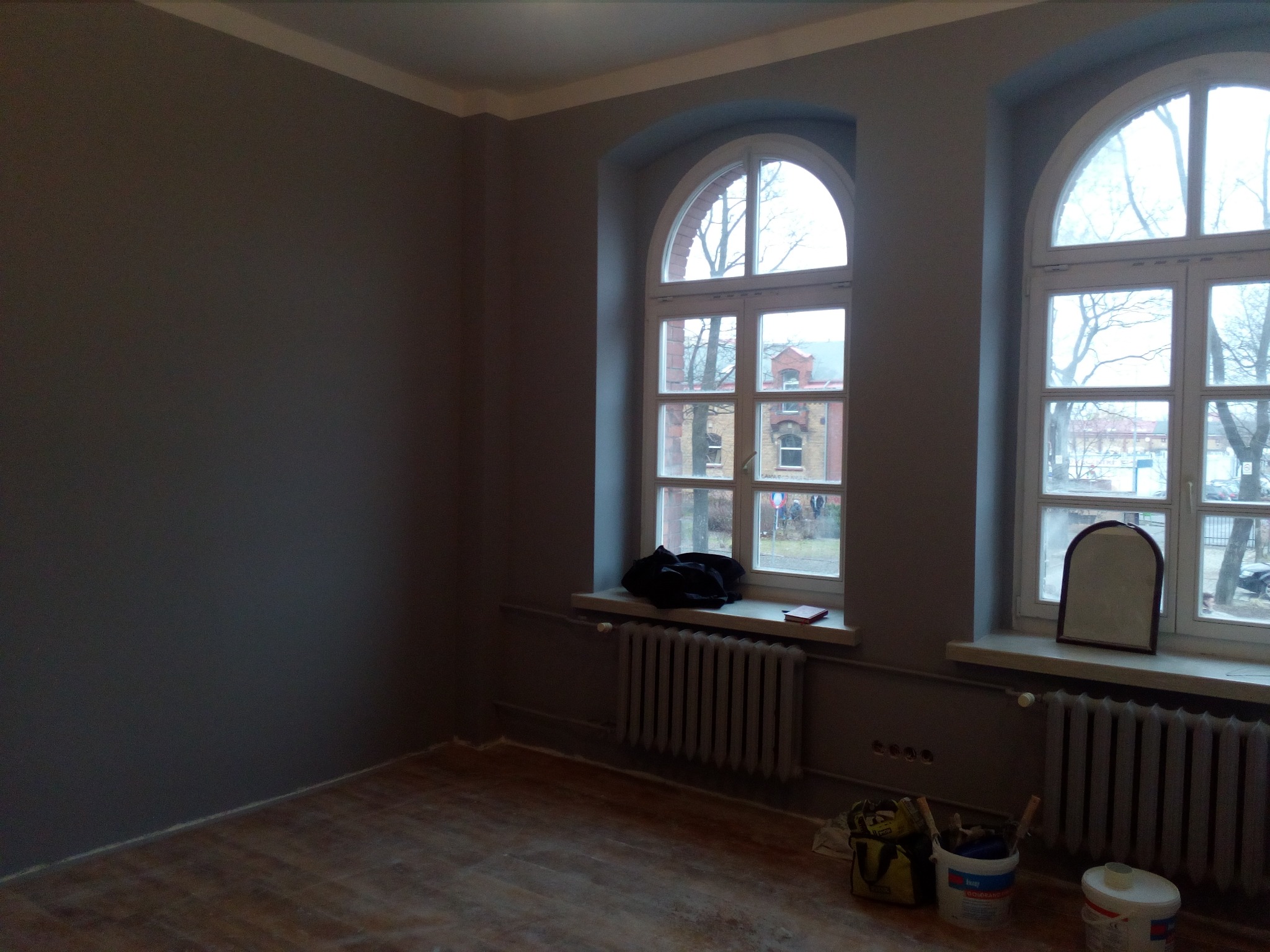 Wnętrze pokoju po malowaniu ścian na szaro, z widocznymi dwoma oknami łukowymi, grzejnikami i wiaderkami z farbą na podłodze.