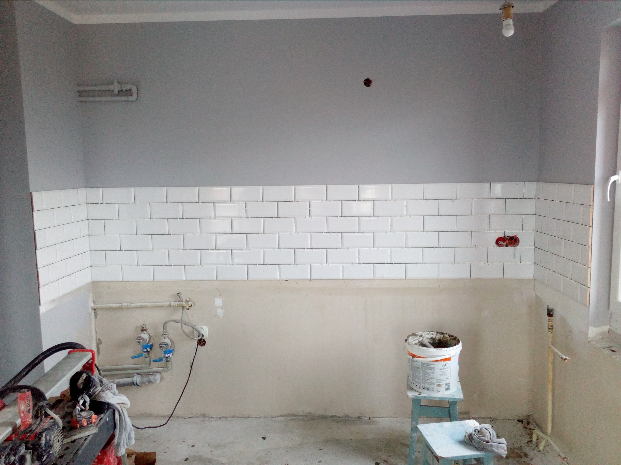 Remont kuchni w trakcie: częściowo wyłożone białe kafelki na ścianie, widoczne narzędzia, wiadro z zaprawą na taborecie, szare ściany w górnej części, instalacja hydrauliczna.