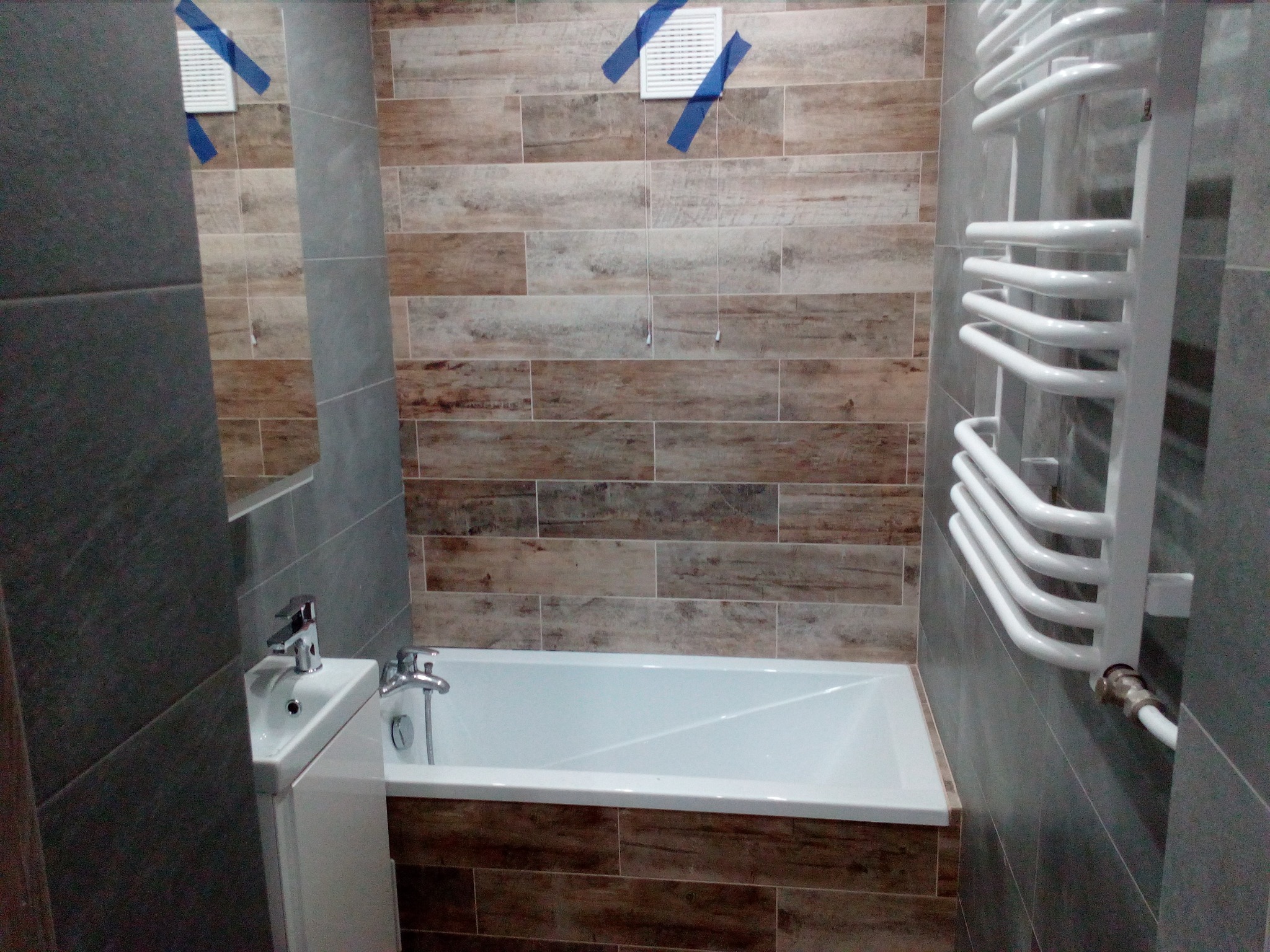 Wykończona łazienka z wanną, umywalką nablatową i grzejnikiem drabinkowym, wyłożona płytkami imitującymi drewno i szarym kamieniem, wentylator przymocowany taśmą malarską.