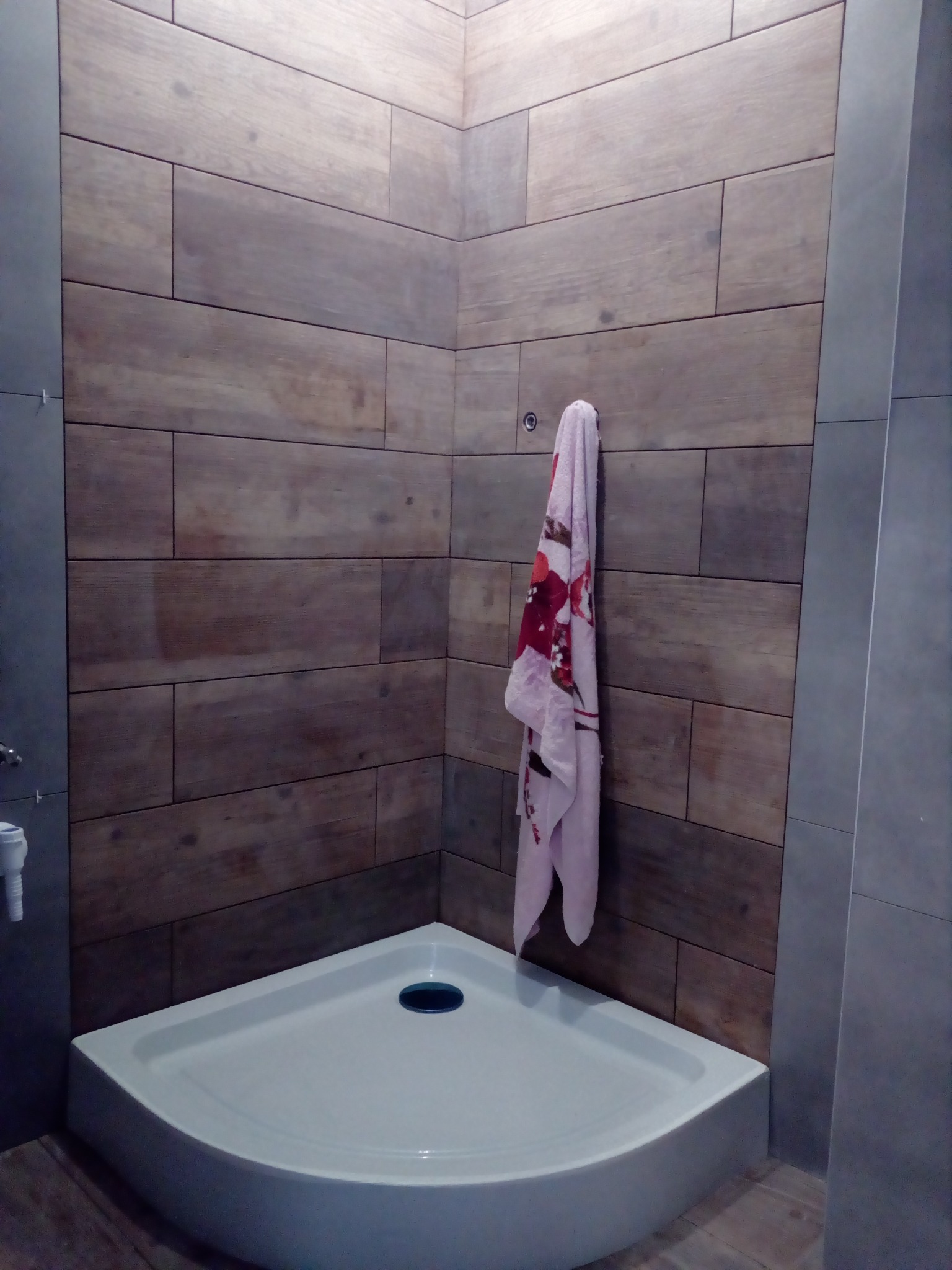 Narożny brodzik z odpływem, ściany wyłożone płytkami imitującymi drewno, wiszący wzorzysty ręcznik, szare płytki na pozostałych ścianach.