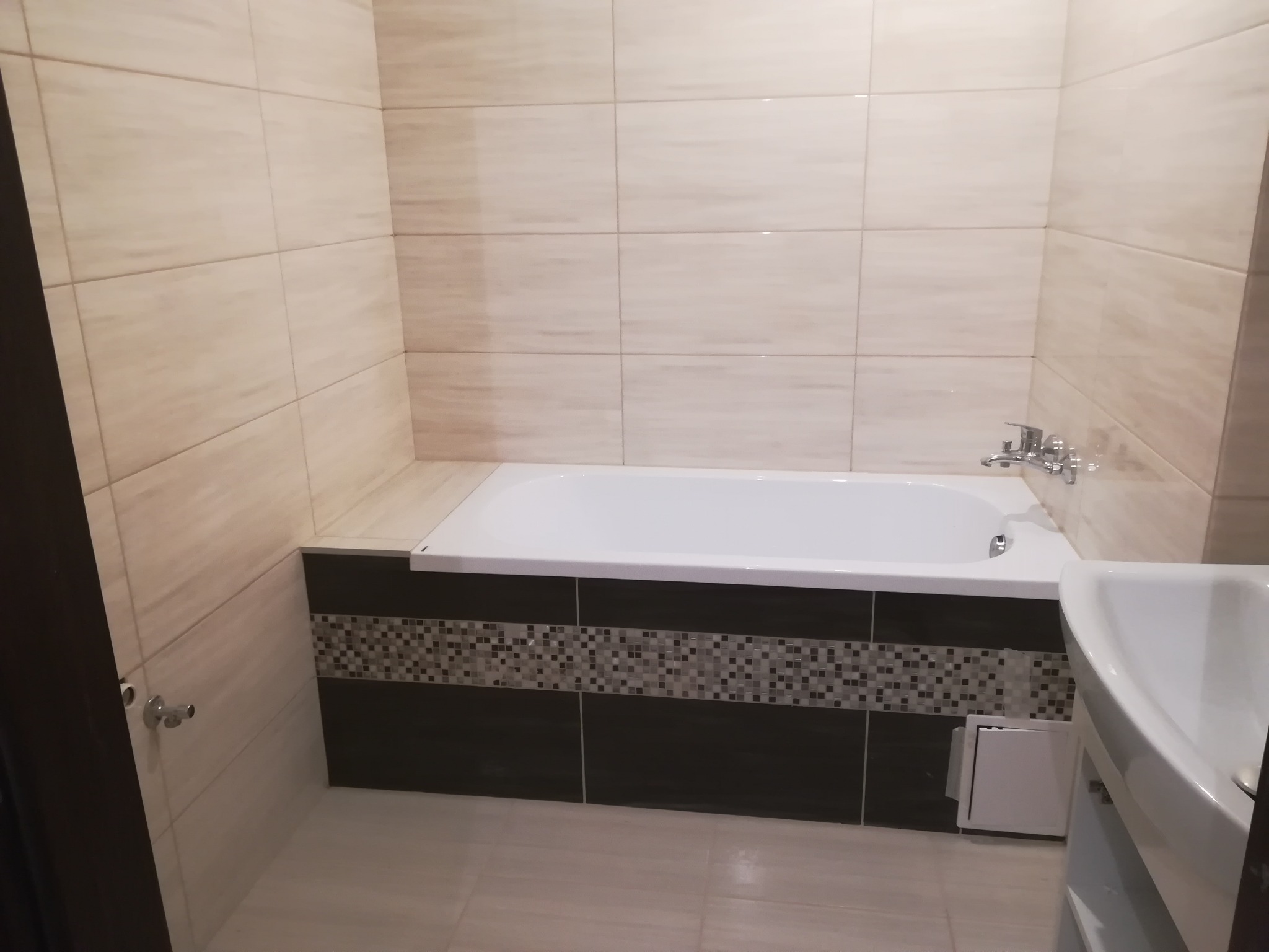 Wykończona łazienka z beżowymi płytkami ściennymi, białą wanną obudowaną ciemnymi płytkami z mozaiką oraz białą umywalką.