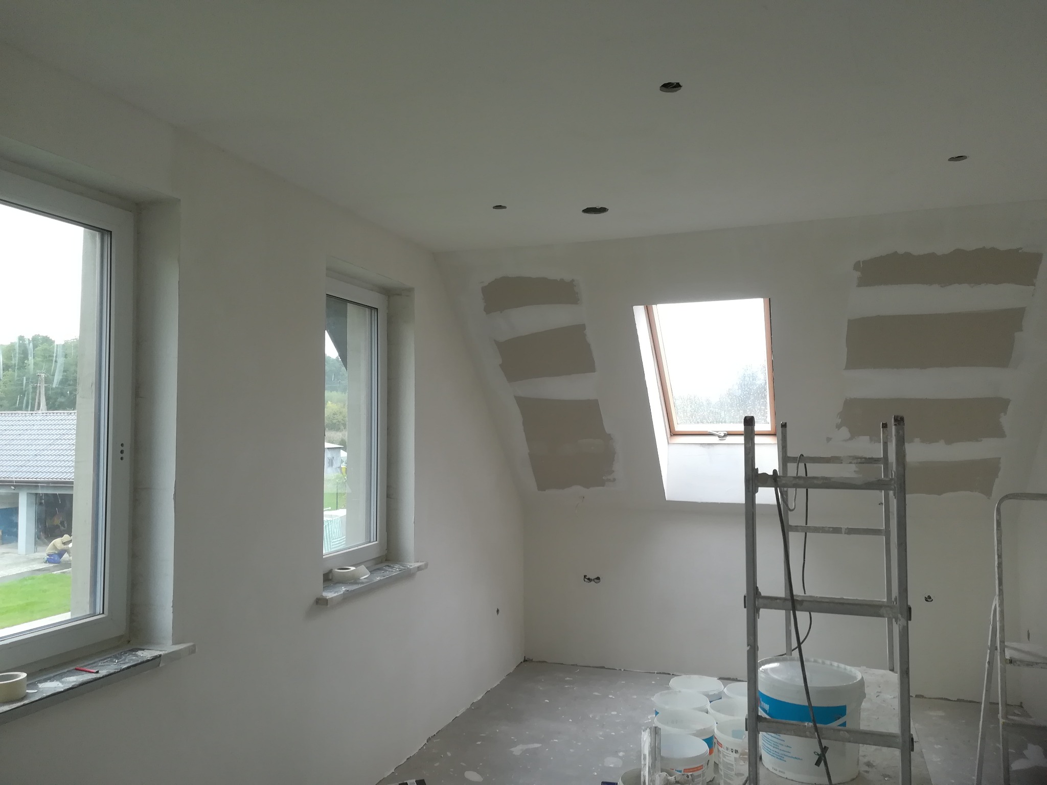 Poddasze po częściowym położeniu gładzi szpachlowej na ścianach, widoczne okna dachowe i pionowe, drabina oraz wiadra z gładzią.