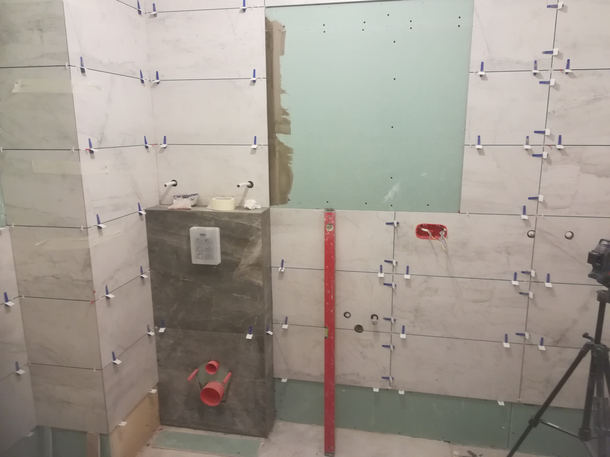 Łazienka w trakcie układania płytek imitujących marmur, widoczny stelaż podtynkowy WC z podłączeniami, poziomica pionowa i system poziomowania płytek z niebieskimi klipsami.