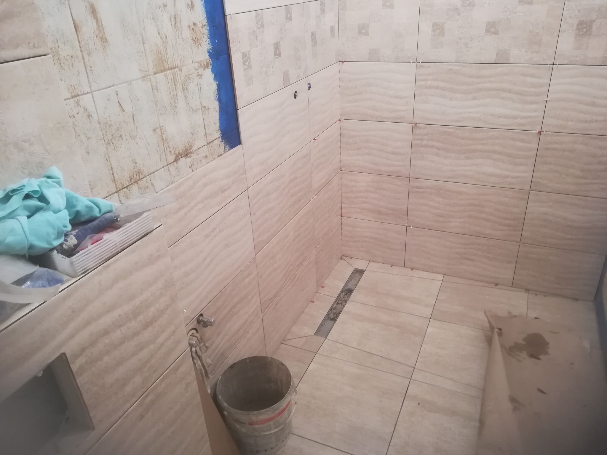 Układanie beżowych płytek imitujących fale w trakcie prac wykończeniowych w łazience, widoczny odpływ liniowy, wiaderko i narzędzia.