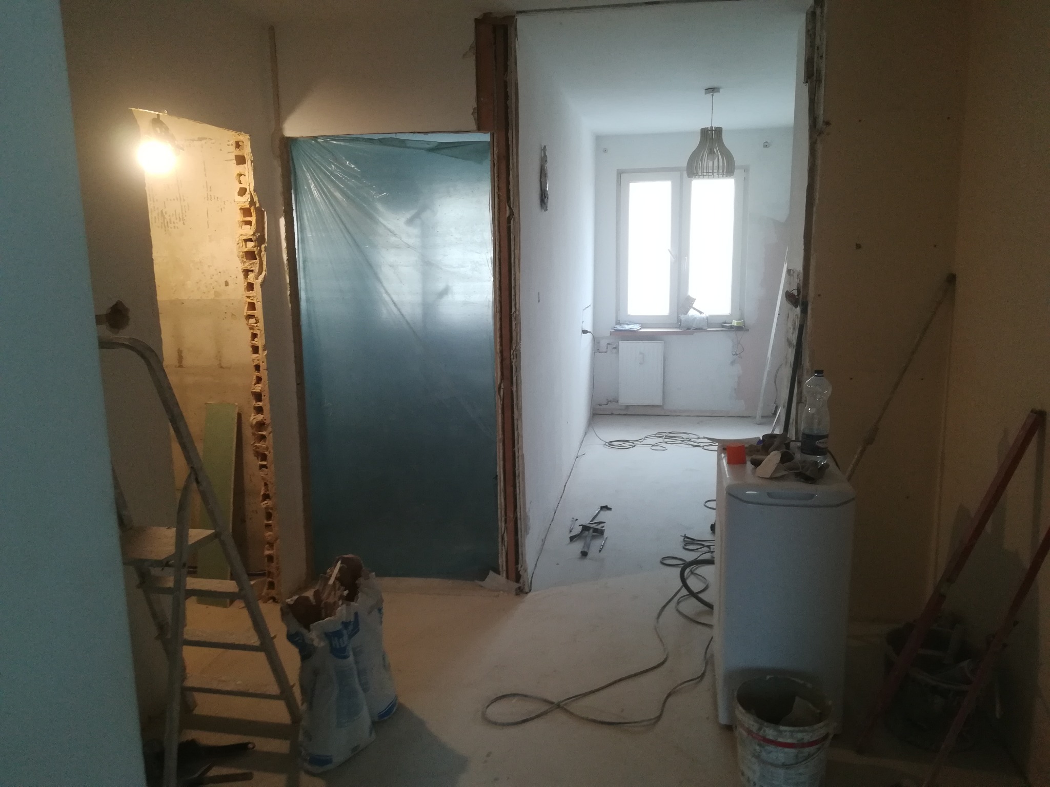 Wnętrze w trakcie remontu: odsłonięta cegła przy futrynie, zabezpieczone folią drzwi, drabina, worki z materiałami budowlanymi, sprzęt AGD i narzędzia w pomieszczeniu z oknem.