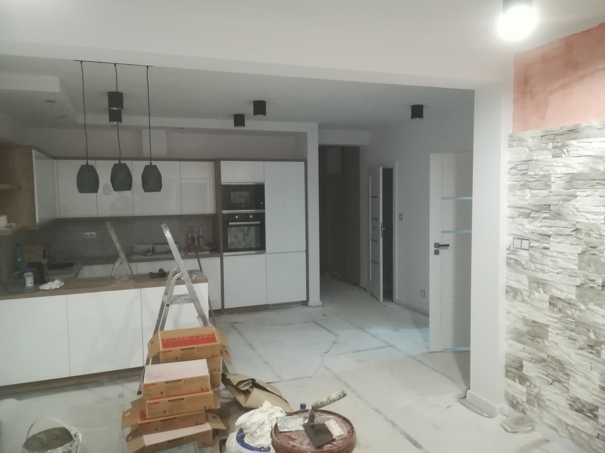 Wnętrze w trakcie wykończenia: biała kuchnia z wyspą i czarnymi lampami, ściana z dekoracyjną okładziną imitującą kamień, widoczne narzędzia i materiały budowlane, drabina oparta o blat.
