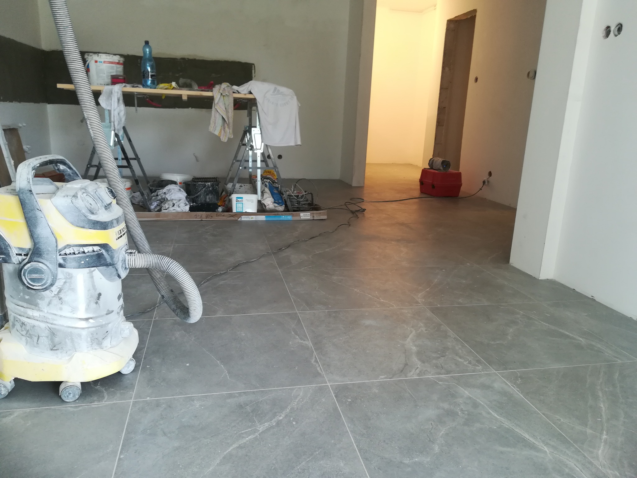 Wnętrze w trakcie remontu: szary gres na podłodze, drabina z narzędziami, odkurzacz przemysłowy i ściany przygotowane do malowania.