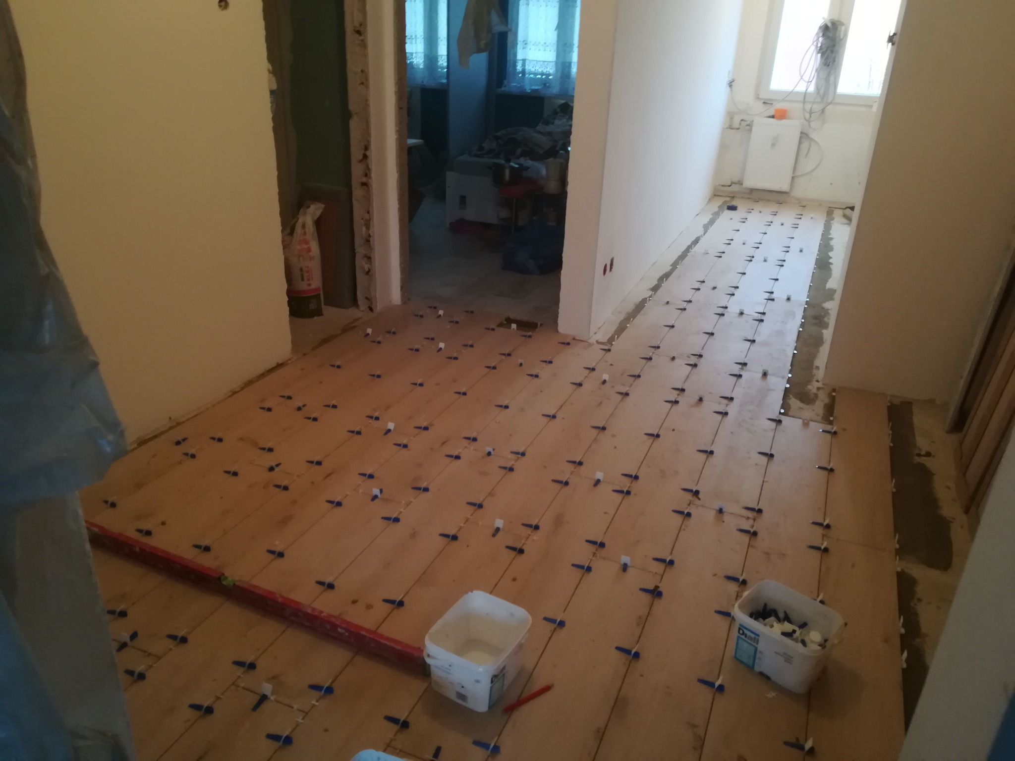 Przygotowanie do układania płytek na podłodze: drewniana podłoga z rozmieszczonymi niebieskimi krzyżykami dystansowymi, poziomica i pojemniki z elementami do poziomowania płytek.