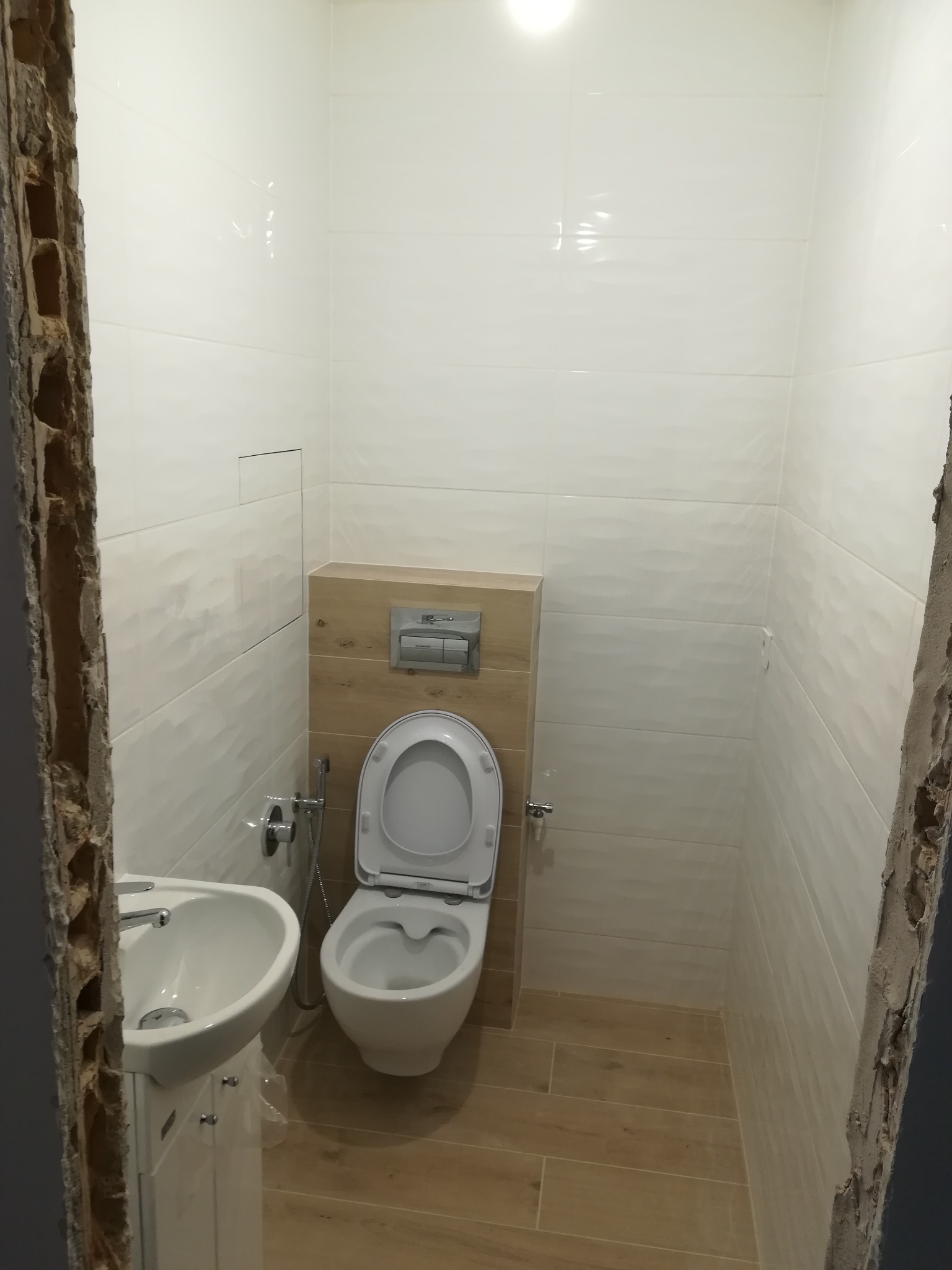 Wykończona łazienka z białymi płytkami o falującej strukturze, drewnopodobną zabudową toalety i małą umywalką na szafce, widoczne surowe wykończenie ościeżnicy.