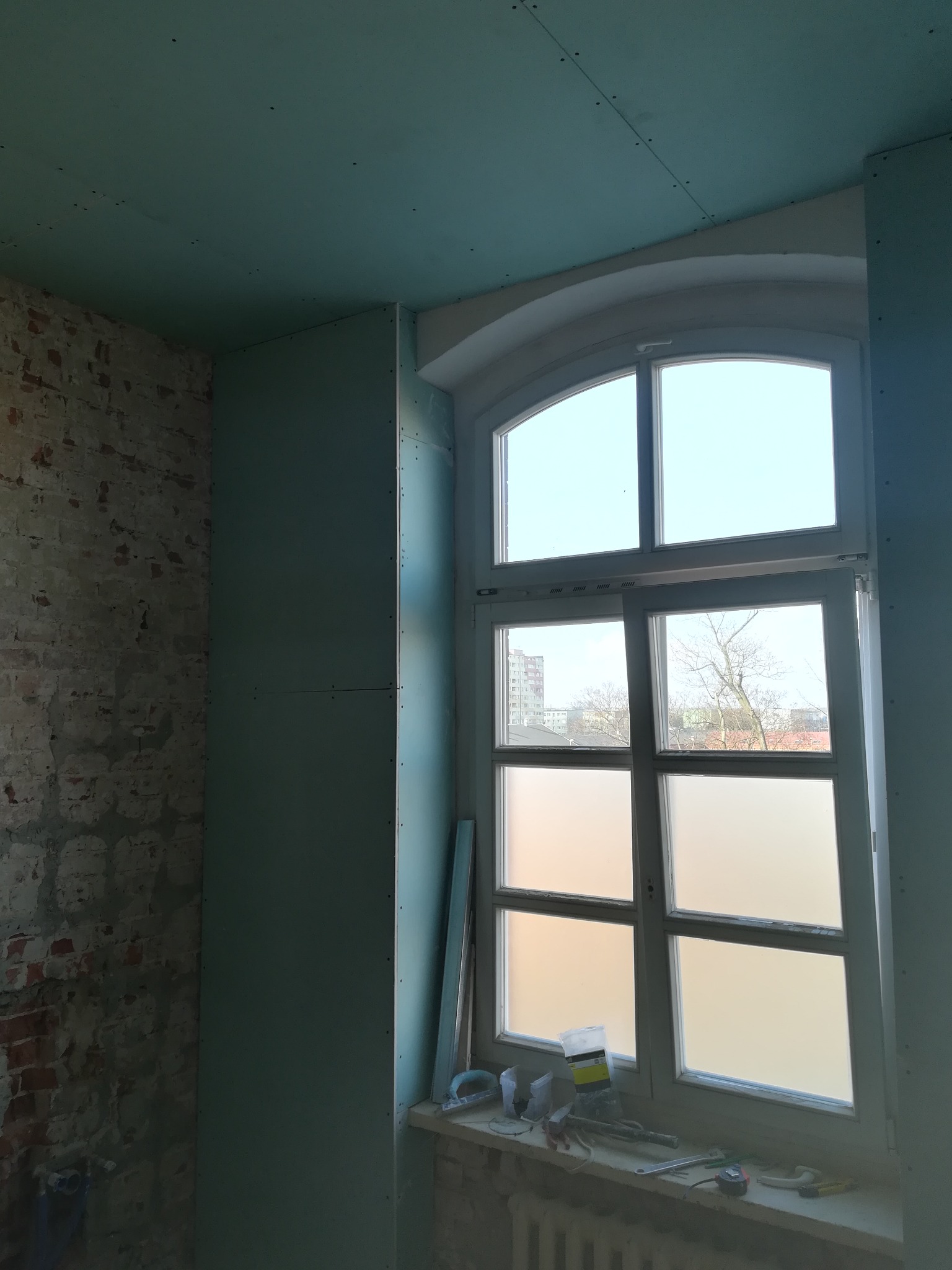 Montaż płyt gipsowo-kartonowych na ścianach i suficie w pokoju z oknem o łukowatym kształcie, widoczny fragment surowej ceglanej ściany i narzędzia na parapecie.