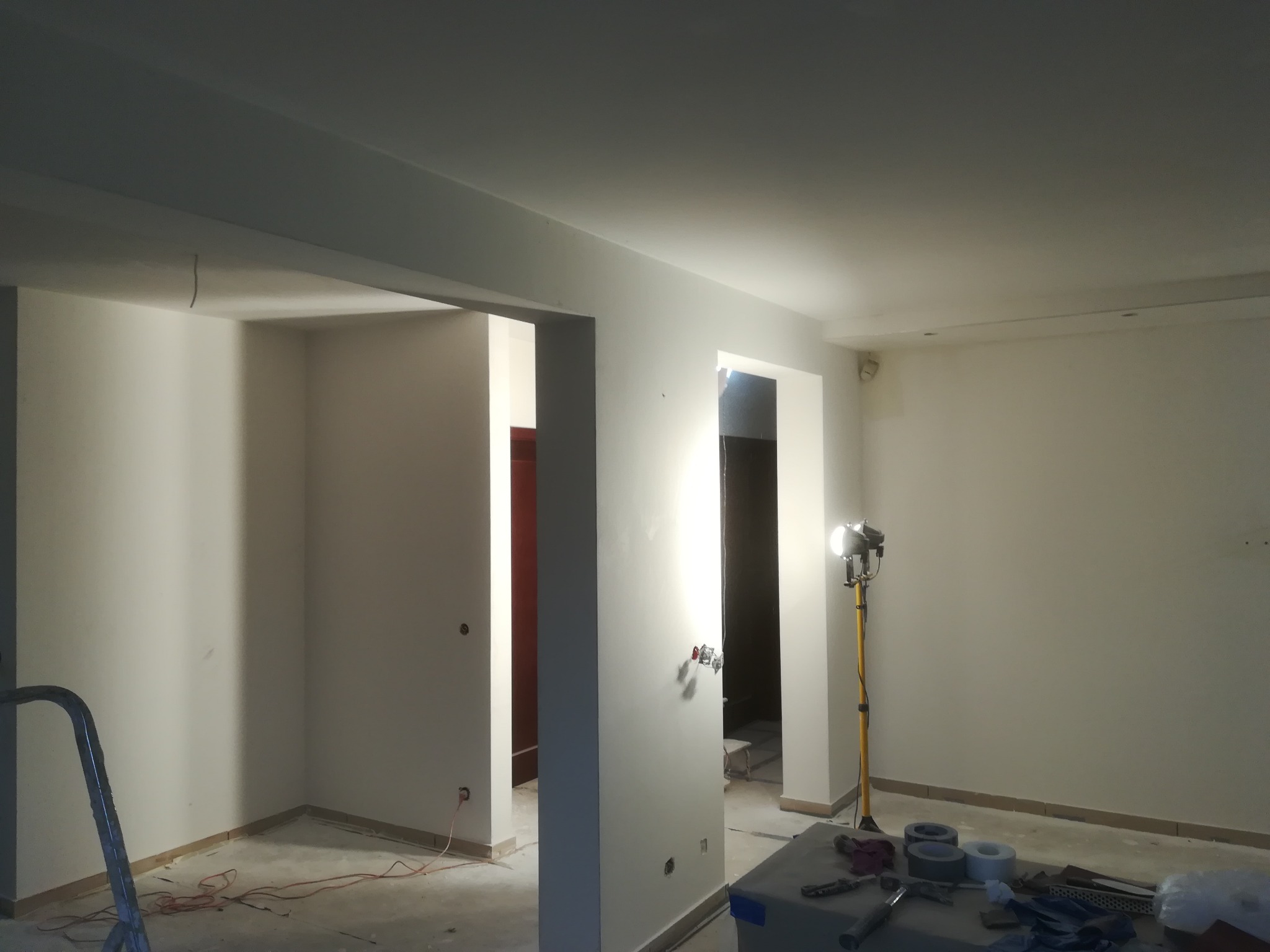 Wnętrze w trakcie prac wykończeniowych: świeżo pomalowane ściany na biało, widoczna drabina, lampa halogenowa na statywie, narzędzia na stole, przygotowane do dalszych prac. Widoczne gniazdka...