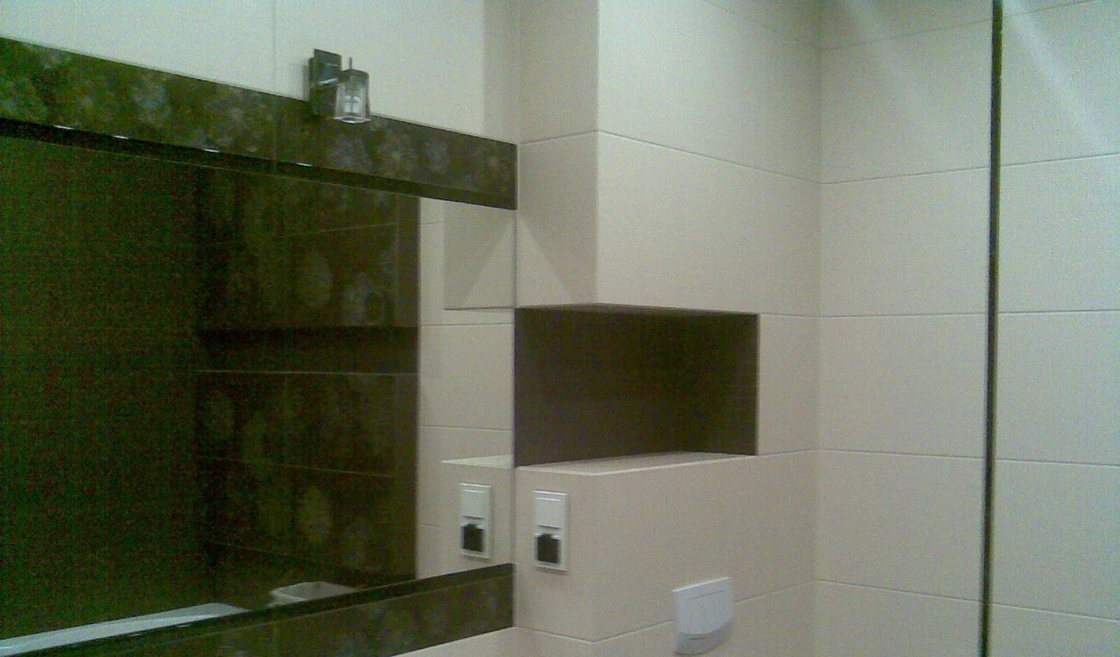 Wykończona łazienka z beżowymi i ciemnobrązowymi płytkami, lustro z ciemną ramą i oświetleniem punktowym, wbudowana półka i białe gniazdka elektryczne.