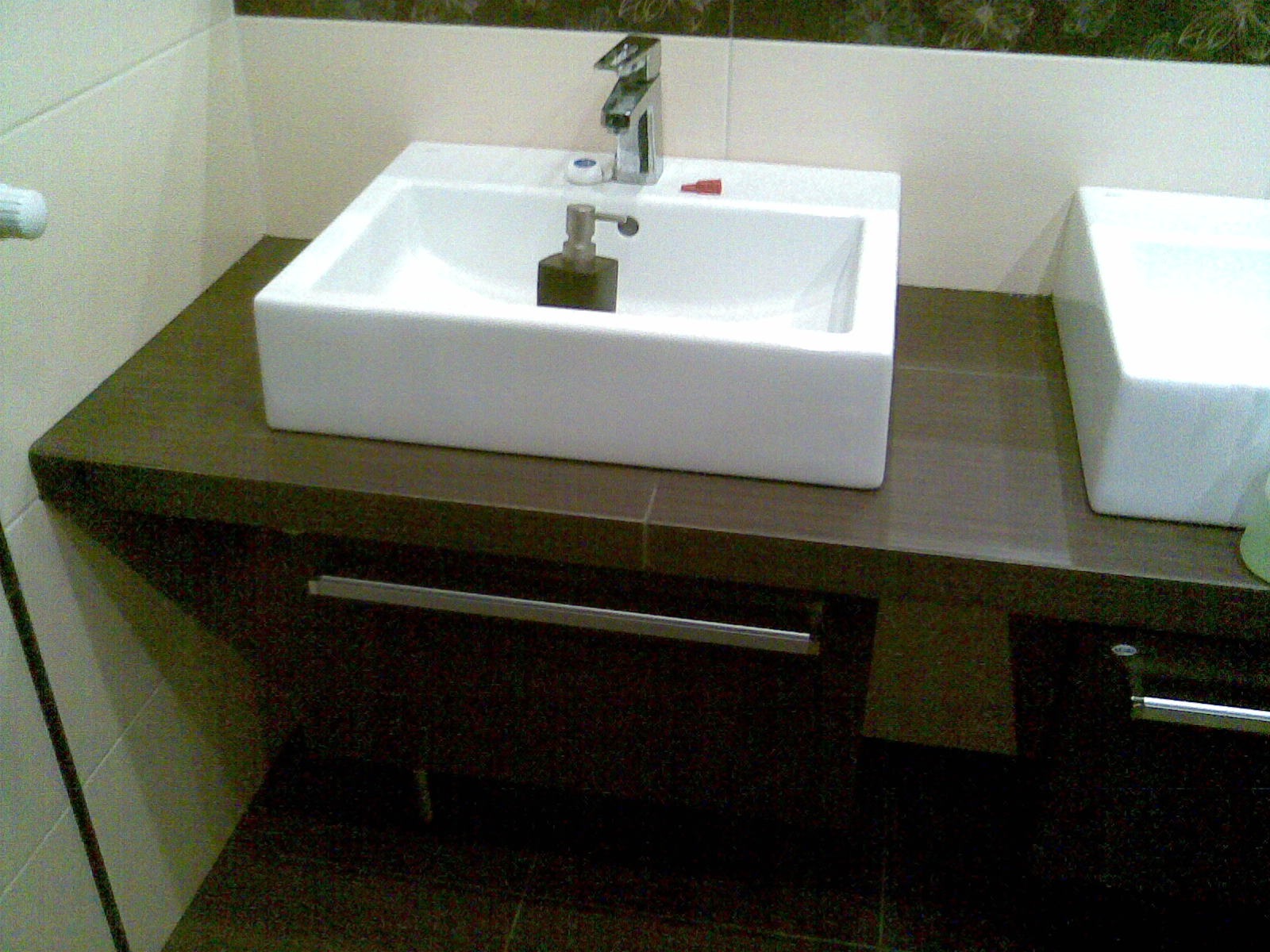 Dwie białe, prostokątne umywalki nablatowe z chromowanymi bateriami, zamontowane na ciemnobrązowym blacie z płytek, nad szafkami z metalowymi uchwytami, w wykończonej łazience.