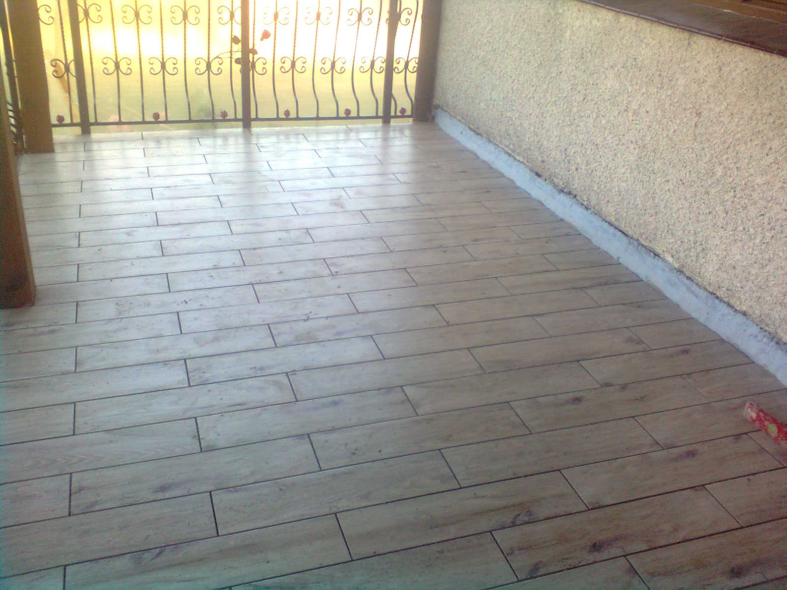 Ułożona podłoga z płytek imitujących drewno na balkonie, widoczna struktura ściany i metalowa balustrada z ozdobnymi elementami.