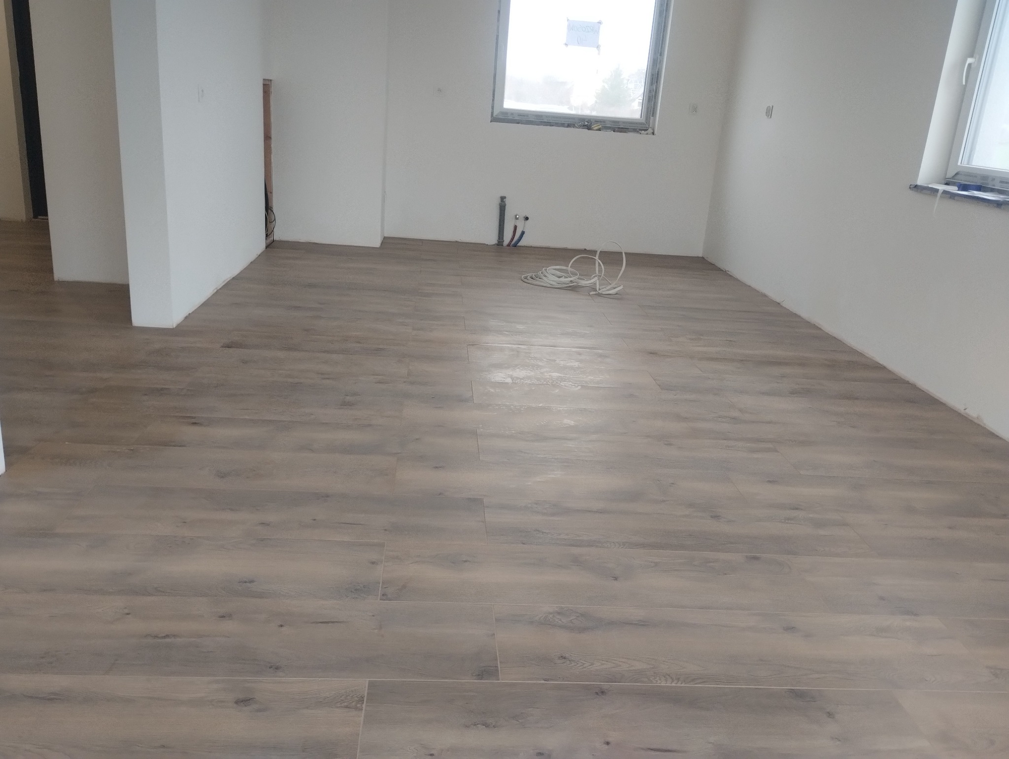 Ukończona podłoga z paneli w odcieniach szarości w jasnym, odnowionym pokoju. Widoczne białe ściany, okno i wystające rury. Kabel leży na podłodze.
