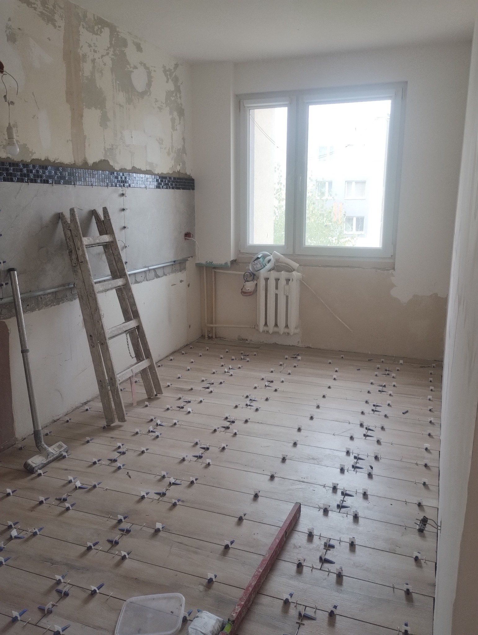 Układanie płytek podłogowych w jasnym wnętrzu. Widoczne krzyżyki dystansowe, drabina oparta o ścianę z fragmentem mozaiki. Okno w tle.