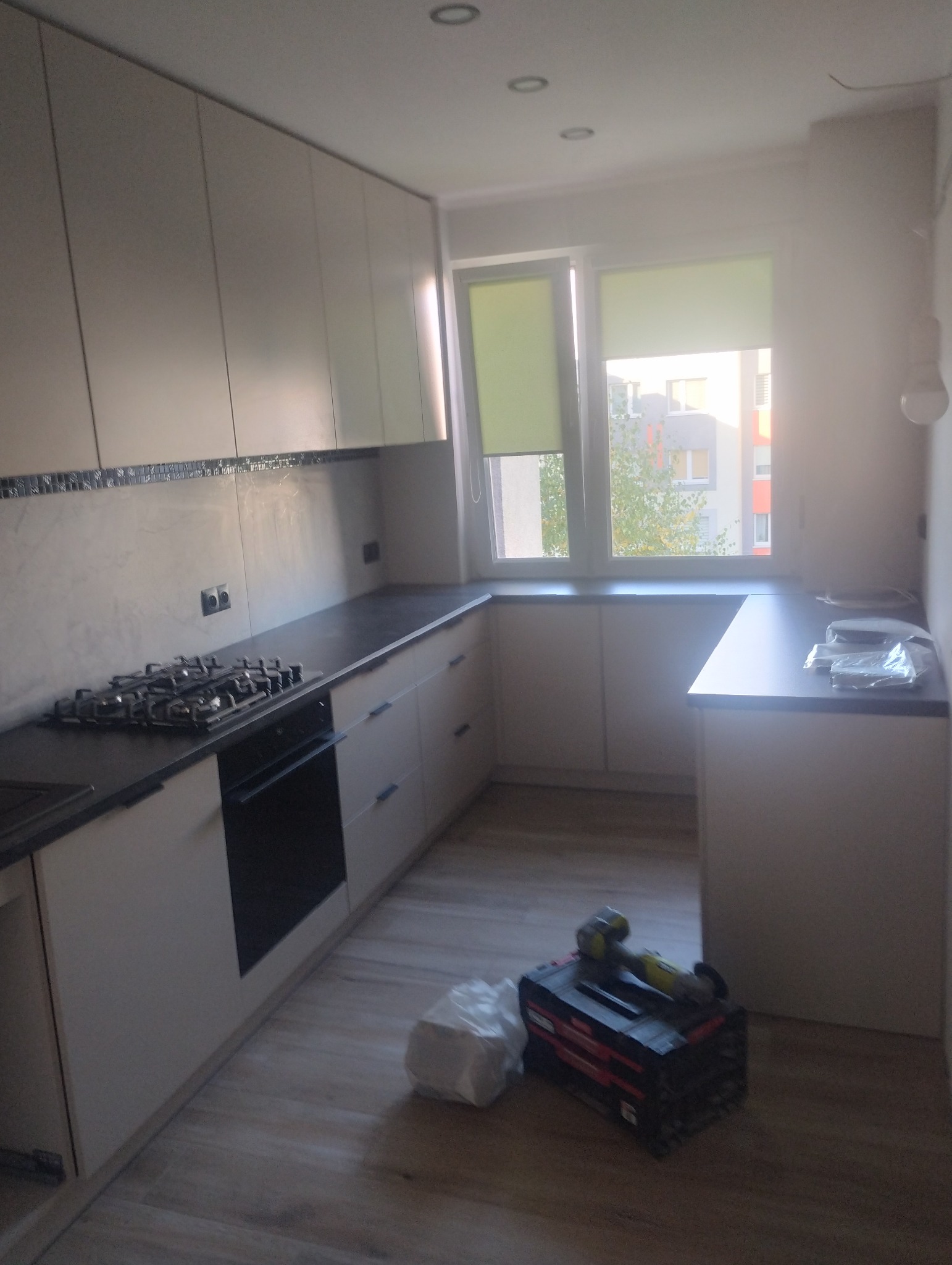 Jasna kuchnia po remoncie z drewnianą podłogą. Widoczne szafki, okno z roletami i narzędzia na podłodze. Minimalistyczny design.