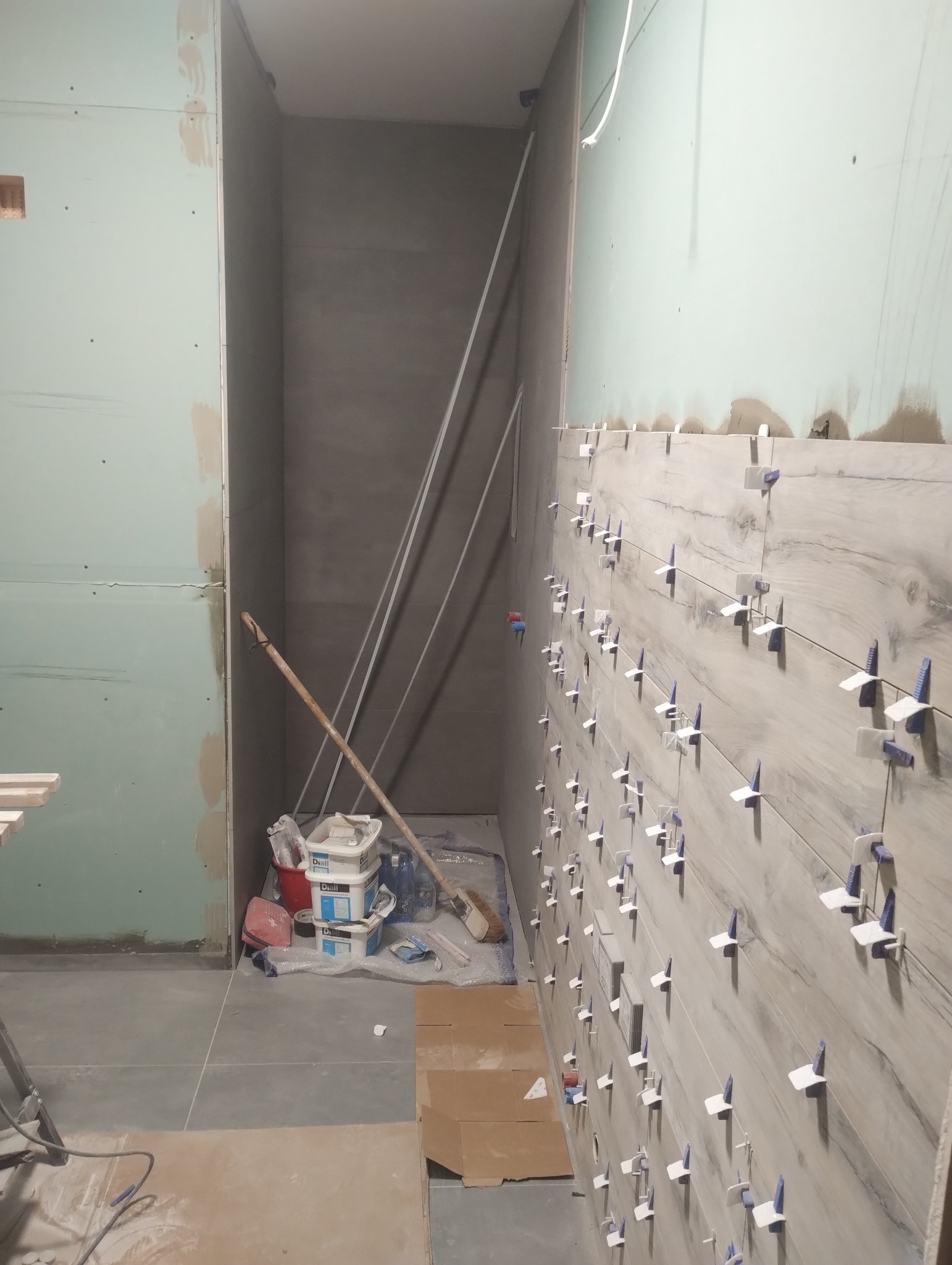 Układanie płytek imitujących drewno na ścianie w łazience. Widoczne krzyżyki dystansowe, narzędzia i materiały budowlane. Ściany przygotowane pod dalsze prace wykończeniowe.