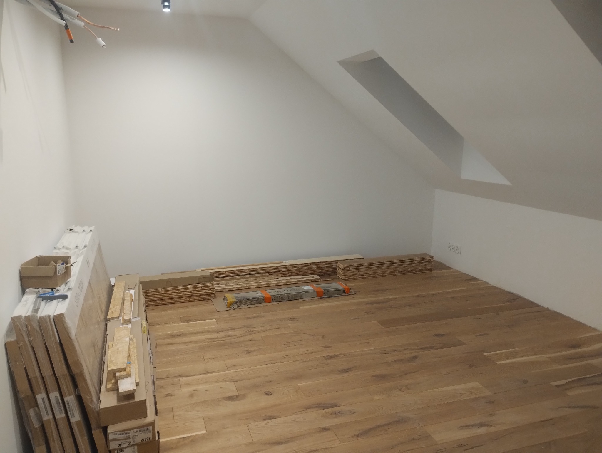 Poddasze z ułożonym parkietem w ciepłym odcieniu drewna. Część paneli w paczkach i rozłożonych, gotowych do montażu. Skosy, okno dachowe, surowy stan wykończenia ścian.